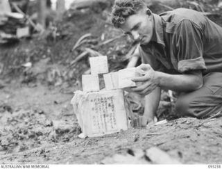 WEWAK AREA, NEW GUINEA, 1945-06-17. PTE R.G. BERESFORD, 2/4 INFANTRY BATTALION, EXAMINING SOME OF THE 4,000 URNS FOUND IN A JAPANESE SHRINE ON THE KOIGIN TRACK; THE URNS, CUBE SHAPED AND MEASURING ..