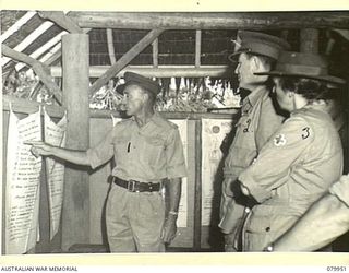 LAE, NEW GUINEA. 1945-03-27. NGX373 MAJOR H.L.R. NIALL (1) AT THE NATIVE MEDICAL SCHOOL, TRANSLATING A HYGIENE CHART (WRITTEN IN PIDGIN ENGLISH) DURING A TOUR OF THE AREA BY LORD WAKEHURST, KCMG, ..