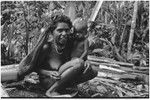 Woman with netbag holds infant, firewood stacked nearby