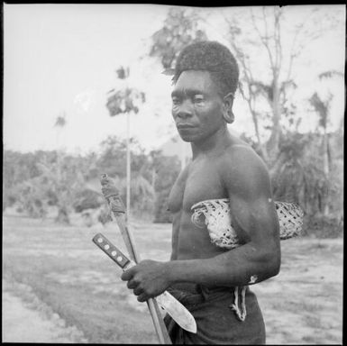 Man holding a knife and a carved stick in one hand with a woven basket under his arm, New Guinea, ca. 1936 / Sarah Chinnery