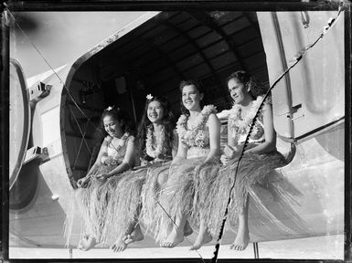 Four unidentified local girls in hula skirts sitting in cargo doorway of C47 transport aircraft, Rarotonga airfield, Cook Islands