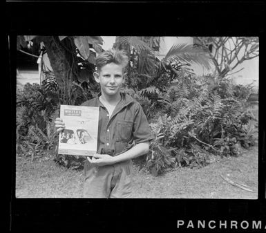 Portrait of an unidentified older boy holding a copy of Whites Aviation magazine Vol 1 No 6 for August 1945 in front of ferns, Rarotonga, Cook Islands