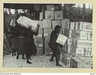 JACQUINOT BAY, NEW BRITAIN, 1945-04-28. A LINE OF NATIVE BOYS CARRYING PARCELS OF SWEETS INTO THE AUSTRALIAN COMFORTS FUND STORE