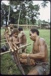 Canoe-building: men use lengths of vine to attach an outrigger float to a canoe, young child observes