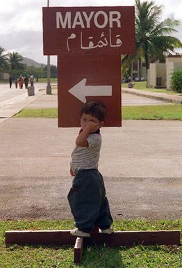 An evacuated Kurdish boy stands next to a sign pointing the way to the "Mayor's" office at Andersen Air Force Base, Guam, during OPERATION PACIFIC HAVEN. Pacific Haven airlifted of over 2,100 Kurds from northern Iraq. The evacuees will be housed temporarily at Andersen AFB, Guam, while they go through the immigration process for residence into the United States
