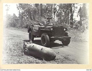 CAPE PUS, WEWAK AREA, NEW GUINEA, 1945-09-07. DRIVER D. COOMBS, HEADQUARTERS 6 DIVISION, IN A JEEP, PASSING AN UNEXPLODED HEAVY TYPE, JAPANESE AERIAL BOMB LYING ON THE ROAD TO CAPE PUS