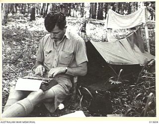 1942-11-19. NEW GUINEA. KOKODA. WAR CORRESPONDENT TOM FAIRHALL OF THE SYDNEY "TELEGRAPH". BESIDE HIM IS A JAPANESE WATER BOTTLE. IN THE BACKGROUND ARE HIS SPACIOUS SLEEPING QUARTERS, KOKODA TRACK ..