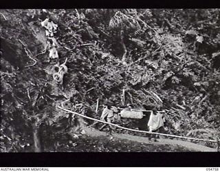 BULLDOG ROAD, NEW GUINEA. 1943-07-19. NATIVES CARRYING COMPRESSOR PARTS ACROSS A LOG BRIDGE AT ECCLESTONE GAP