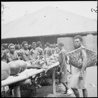 Man with arm through a bag, in front of water tanks, Boong, native market, Rabaul, New Guinea, ca. 1936 / Sarah Chinnery