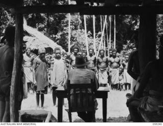 WIDERU, NEW GUINEA, 1943-10-21. NX155085 CAPTAIN R.G. ORMSBY OF THE AUSTRALIAN AND NEW GUINEA ADMINISTRATIVE UNIT SITTING AT HIS DESK, ADDRESSING THE VILLAGE NATIVES IN A TALK ON THE WAR AND ..