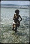 Girl in short fiber skirt walks in shallow lagoon, edge of coral reef visible in distance