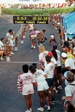 A cameraman records the event with a videocamera as a contestant prepares to cross the finish line after completing the 26-mile marathon, part of the 1987 Ironman Competition