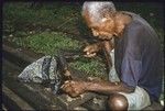 Carving: older man uses an adze with traditional handle and metal blade to shape a piece of wood