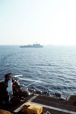 A starboard view of the dock landing ship USS GUNSTON HALL (LSD-44) underway, taken from the superstructure of the amphibious assault ship USS GUAM (LPH-9). Flight deck crewmen service an SH-2F Seasprite helicopter on the GUAM. The amphibious transport dock USS AUSTIN (LPD-4) and the tank landing ships USS LA MOURE COUNTY (LST-1194) and USS SUMTER (LST-1181) are in the background
