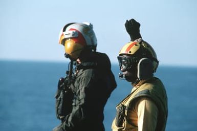 A helicopter launch petty officer directs flight deck operations aboard the amphibious assault ship USS GUAM (LPH 9) during operations off the coast of Lebanon. A pilot passes by in the background