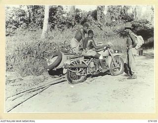 HANSA BAY, NEW GUINEA. 1944-06-16. SERGEANT T. RAFTY, MILITARY HISTORY SECTION, WATCHING TWO NATIVE LADS TRYING TO START THE MOTOR OF AN ABANDONED JAPANESE MOTOR CYCLE