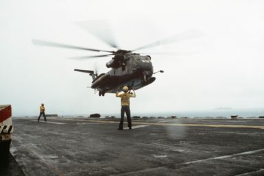 A plane director signals instructions to the pilot of a Helicopter Mine Countermeasures Squadron 14 (HM-14) RH-53D Sea Stallion helicopter hovering over the flight deck of the amphibious assault ship USS GUAM (LPH 9)