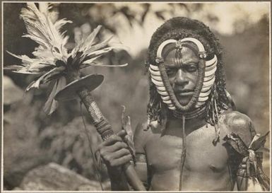 [Man wearing a mask and carrying a club with many feathers at the top] Frank Hurley