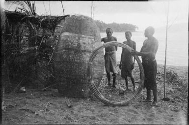 Two men standing beside a fishing basket and one man holding a bound coil of cane, New Guinea, ca. 1929 / Sarah Chinnery