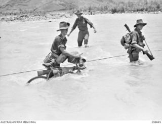 RAMU VALLEY, NEW GUINEA, 1943-10-05. A RUNNER OF "A" COMPANY, 2/27TH AUSTRALIAN INFANTRY BATTALION WHEELING HIS CYCLE LADEN WITH RIFLES AND OTHER GEAR ACROSS THE SURINAM RIVER. SHOWN ARE:- NX127253 ..