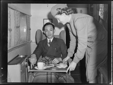 Interior view of aircraft, includes stewardess in uniform serving meal to unidentified man on Tasman Empire Airways air boat RMA New Zealand ZK-AME, flight to Fiji