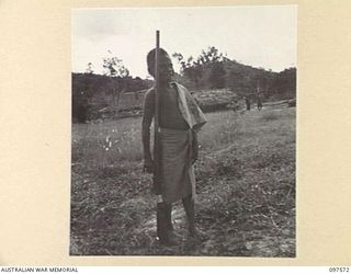 MAPRIK, NEW GUINEA. 1945-10-03. AN AUSTRALIAN NEW GUINEA ADMINISTRATIVE UNIT NATIVE BOY AT AUSTRALIAN NEW GUINEA ADMINISTRATIVE UNIT HEADQUARTERS STANDING AT THE "ORDER" WITH A SHOT GUN BIGGER THAN ..