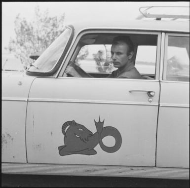 A man behind the steering wheel of a car with an image of a capricorn on the door, New Caledonia, 1967 / Michael Terry