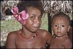 Adolescent girl, hibiscus flower in hair and dabs of betel nut paste on cheeks, holds an infant, both wear shell necklaces