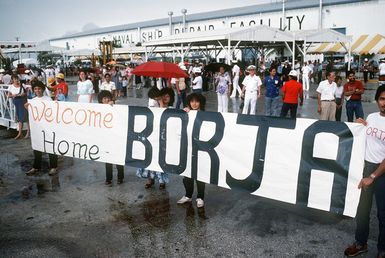 Family members display a welcome home banner for a sailor aboard the combat stores ship USS NIAGARA FALLS (AFS 3). The ship is arriving at Naval Station Guam