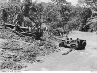 FINSCHHAFEN AREA, NEW GUINEA. 19 NOVEMBER 1943. A BULLDOZER TOWS A JEEP CARRYING SUPPLIES IN BOXES AND TINS ACROSS THE QUOJA RIVER AT A CROSSING ON THE MAIN TRACK TO THE FORWARD AREAS FROM ..