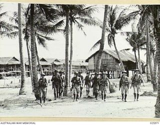 PORT MORESBY, PAPUA. 1942-07-20. AUSTRALIAN SOLDIERS RETURNING FROM PATROL, MOVING THROUGH COCONUT PALMS WITH TYPICAL NATIVE VILLAGE IN THE BACKGROUND