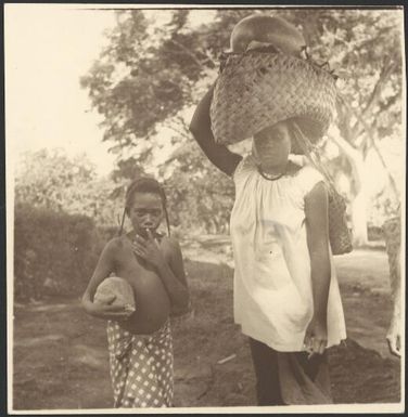 Woman carrying a bag on her head and a child with a coconut walking to the markets, Rabaul, New Guinea, ca. 1936 / Sarah Chinnery