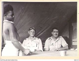 WUNUNG PLANTATION, JACQUINOT BAY, NEW BRITAIN. 1944-12-08. A NATIVE, PRIVATE SIMET, CARRYING A CUP OF TEA TO ALLIED INTELLIGENCE BUREAU OFFICERS SEATED IN A TENT. IDENTIFIED PERSONNEL ARE:- FLIGHT ..