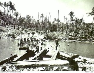 NEW IRELAND, 1945-10. NATIVE CARRIERS USING A DAMAGED BRIDGE TO CROSS A RIVER. (RNZAF OFFICIAL PHOTOGRAPH.)
