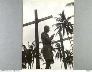 1942-12-16. GONA, PAPUA. CHURCH OF ENGLAND PADRE A.E. BEGBIE IN A CEMETERY BETWEEN TWO CROSSES MARKING THE GRAVES OF SOLDIERS WHO DIED DURING THE BATTLE FOR GONA. (NEGATIVE BY G. SILK)