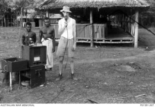 MAPRIK? NEW GUINEA. C. 1945. SERGEANT N. (NAT) F. DANIELL, A MEMBER OF THE FRONT LINE BROADCASTING UNIT, FAR EASTERN LIAISON OFFICE, WATCHED BY TWO NATIVES, USES A MICROPHONE AND BROADCASTING ..