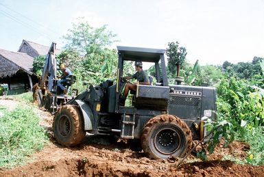 An island resident sits at the operator's station on an excavator as a Seabee from Construction Battalion Unit 3 (CBU-3) backs the vehicle around a hole. Seabees from CBU-3 are working with island residents to make improvements on a school as part of a civic action proejct