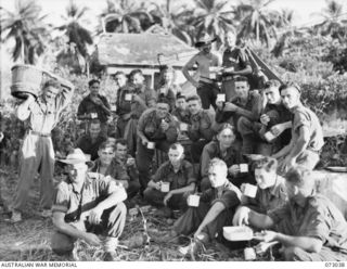 MADANG, NEW GUINEA. 1944-04-28. MEMBERS OF HEADQUARTERS 8TH INFANTRY BRIGADE RELAXING DURING A MEAL. THE RUINS OF THE OLD HOTEL MADANG STAND IN THE BACKGROUND