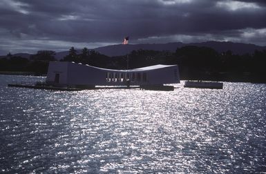 The American flag flies over the USS ARIZONA (BB-39) Memorial as the sun highlights the surrounding waters