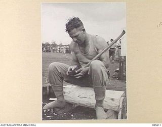 HERBERTON RACECOURSE, WONDECLA, ATHERTON TABLELAND, QLD. 1945-01-19. PRIVATE T WARREN, 2/43 INFANTRY BATTALION SHARPENING HIS AXE PRIOR TO THE WOODCHOPPING EVENT DURING THE 9 DIVISION GYMKHANA