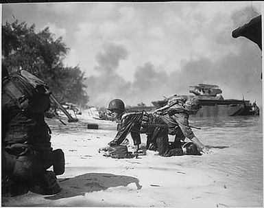 It appears that one Marine is relieving another on the beach at Saipan but they are really crawling under enemy fire, to their assigned positions.