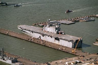 Aerial port quarter view of the amphibious assault ship USS Saipan (LHA-2) tied up on the north side of pier 6. In the upper left of the photo is a Los Angeles class nuclear-powered attack submarine arriving in company of two YTBs