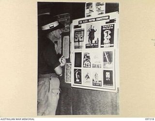 CAPE WOM, NEW GUINEA. 1945-09-29. PRIVATE J. BROMLEY AT HEADQUARTERS 6 DIVISION LOOKING AT ENTRIES IN AN AUSTRALIAN ARMY EDUCATION SERVICE POSTER COMPETITION