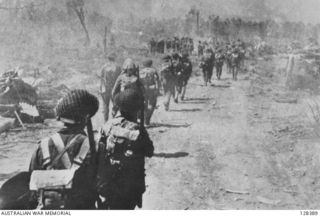 TROOPS OF THE 7TH DIVISION ENTERING LAE ON 1943-09-16 ALONG A ROAD LITTERED WITH DEBRIS FROM AIR AND ARTILLERY BOMBARDMENTS