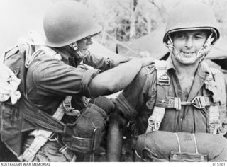 1943-09-15. PARATROOPS LAND AT NADZAB. GUNNER DONNELLY ADJUSTS LANCE-BOMBARDIER WEBB'S PARACHUTE BEFORE THE AUSTRALIAN AIRBORNE ARTILLERYMEN AND THEIR GUNS - DISMANTLED AND ATTACHED TO PARACHUTES - ..