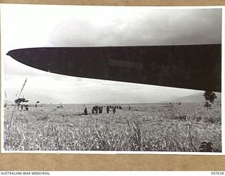 RAMU VALLEY, NEW GUINEA, 1943-10-01. NATIVES CUTTING KUNAI GRASS ON THE RUNWAYS OF THE NEW AIRSTRIP. TRANSPORT AIRCRAFT WING IN THE FOREGROUND