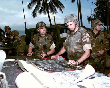 Two US Marines review perimeter security maps at Kaneohe Bay Marine Corps Station, HI during Operation RIMPAC 96. A third Marine stands near a M998 High-Mobility Multipurpose Wheeled Vehicle (HMMWV) while two other Marines use communication equipment in the background