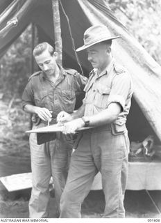 KARAWOP, NEW GUINEA. 1945-05-05. MAJOR B.W.T. CATTERNS, ADMINISTRATIVE COMMANDER, 2/1 INFANTRY BATTALION (1), LISTENS TO THE LATEST INTELLIGENCE INFORMATION INDICATED ON A SITUATION MAP AS POINTED ..