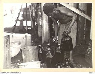 LAE, NEW GUINEA. 1944-12-12. A NATIVE SUPPLIED BY ANGAU WORK UNDER THE SUPERVISION OF CORPORAL A.P. RYAN, ARMY CANTEENS SERVICE, (1), AS THEY LOAD BOTTLES INTO A CLEANING MACHINE AT A SOFT DRINK ..