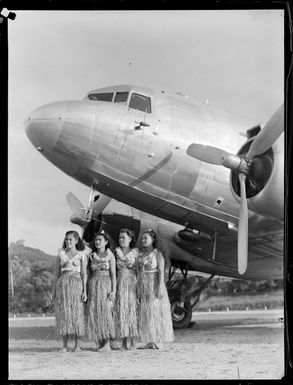 Four unidentified local girls wearing hula skirts in front of a C47 transport aircraft, Rarotonga airfield, Cook Islands